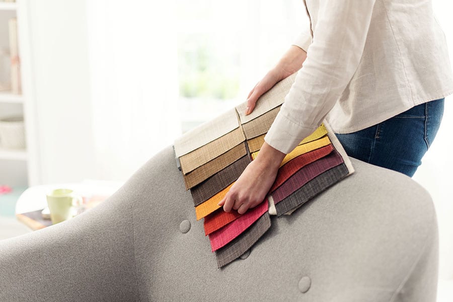 Person in a long sleeve white shirt holding multiple fabric color pieces to a light grey sofa in their home in Springfield, IL