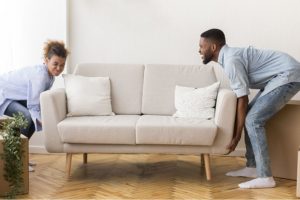 Man and woman in a light blue top and jean bottoms lifting a grey couch and rearranging the furniture in their living room in Springfield, IL.)