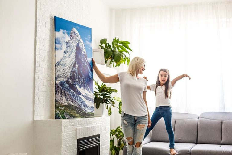 Mother in a white t-shirt and jeans hanging a white, blue, and green mountain canvas with daughter standing on the couch in Petersburg, IL.