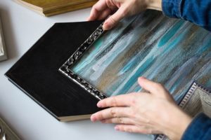 Person in a long sleeve blue shirt choosing a decorative black frame for a blue, white, and black painting for their home in Petersburg, IL.