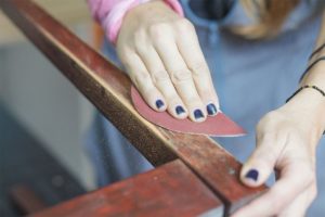 Woman with blue nail polish and a blue apron sanding a piece of vintage furniture and making it modern for her home in Athens, IL.