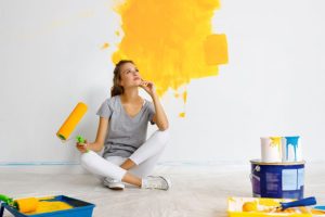 Woman in a grey t-shirt and white pants sitting on the ground with a green paint roller and yellow paint on the walls in Athens, IL.