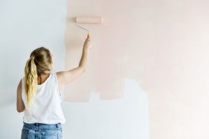 Woman in a white tank top and jean shorts using a rolling brush to paint a blush shade of satin or eggshell paint on her living room wall in Petersburg, IL.
