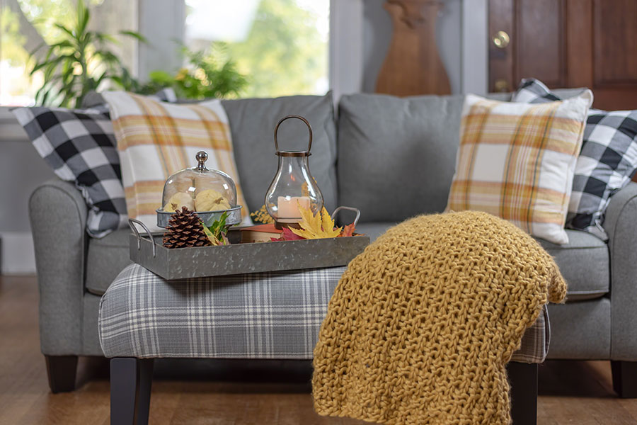A grey reupholstered couch with fall and autumn décors such as checkered pillows and a chunky yellow blanket found at a furniture store in Springfield, IL.